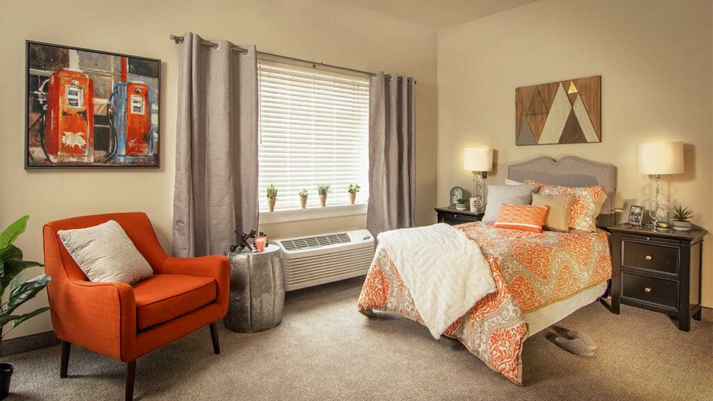 A cozy bedroom with an orange accent chair, a bed with a patterned orange and gray comforter, and two bedside tables with lamps. A large window with blinds is adorned with gray curtains. A colorful painting of old gas pumps hangs on the wall.