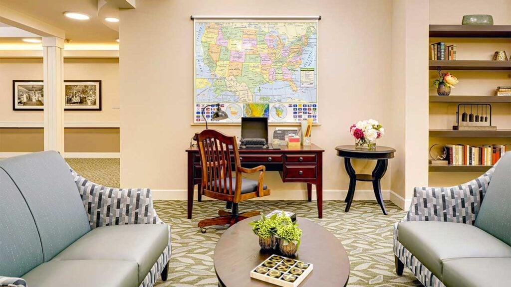 A cozy office with a large map of the USA on the wall, a wooden desk with a chair, a round side table with flowers, and bookshelves. The room has gray couches and a coffee table with plants and decorative items.