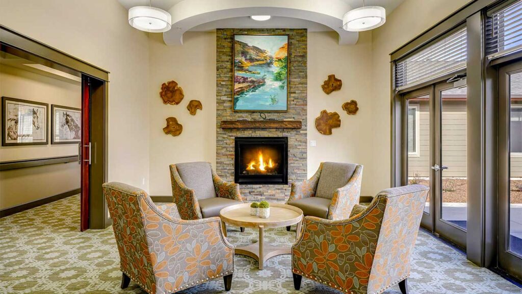 A cozy seating area with four patterned armchairs around a small round table, centered by a stone fireplace. Above the fireplace is a colorful landscape painting. The room is warmly lit with ceiling lights and natural light through windows.