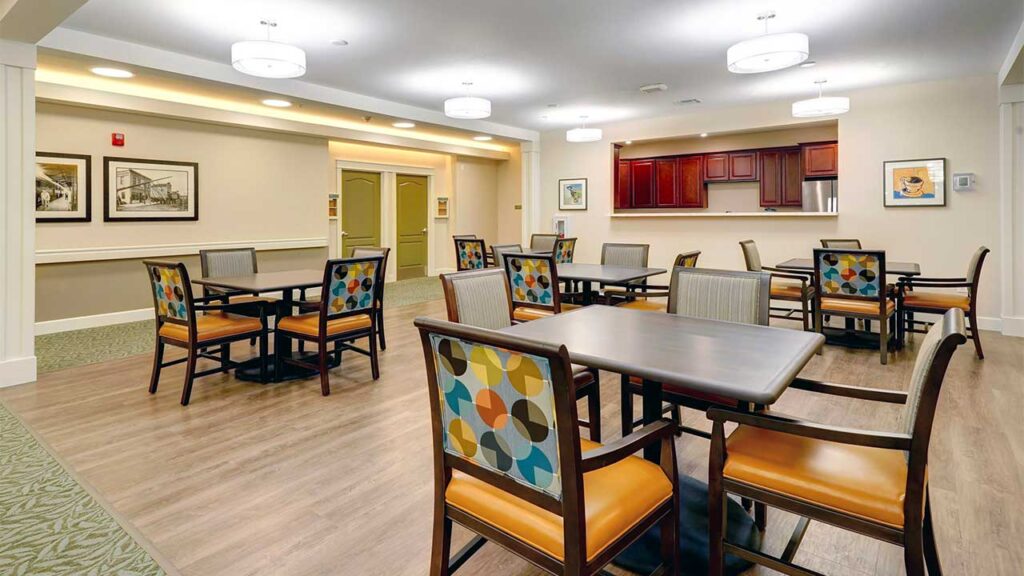 A dining area with wooden tables and chairs featuring colorful back cushions. The room has a warm color scheme, framed artwork on the walls, a service window in the back, and modern overhead lighting fixtures.