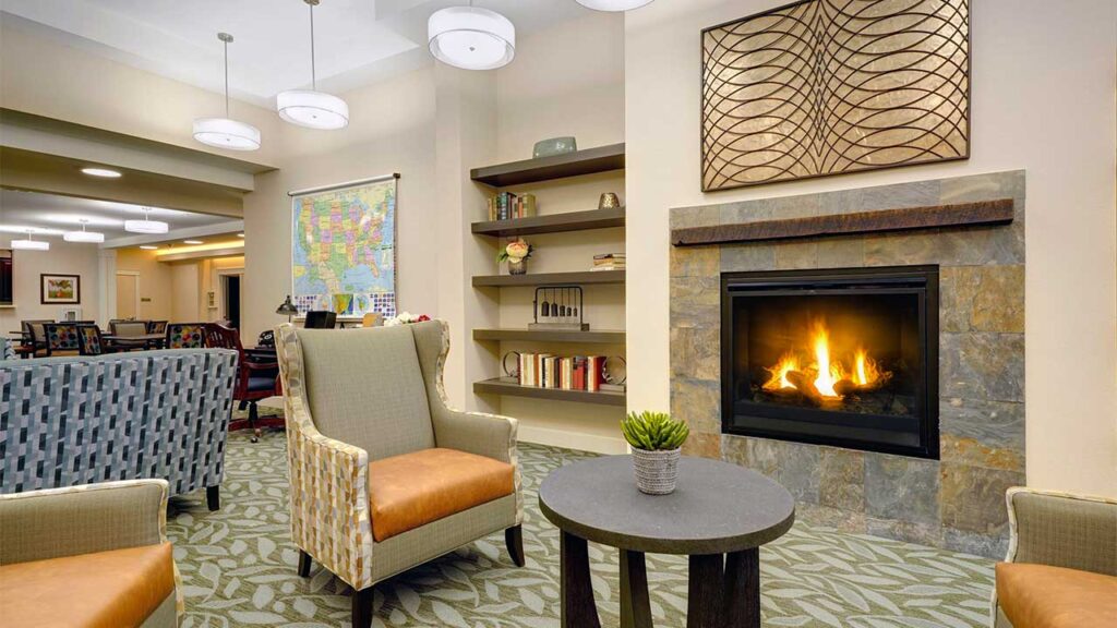 A cozy living room with a lit fireplace, surrounded by armchairs and a round table. Shelves with books and decor adorn the wall. A large map is visible in the background, along with overhead lighting and soft carpeted flooring.