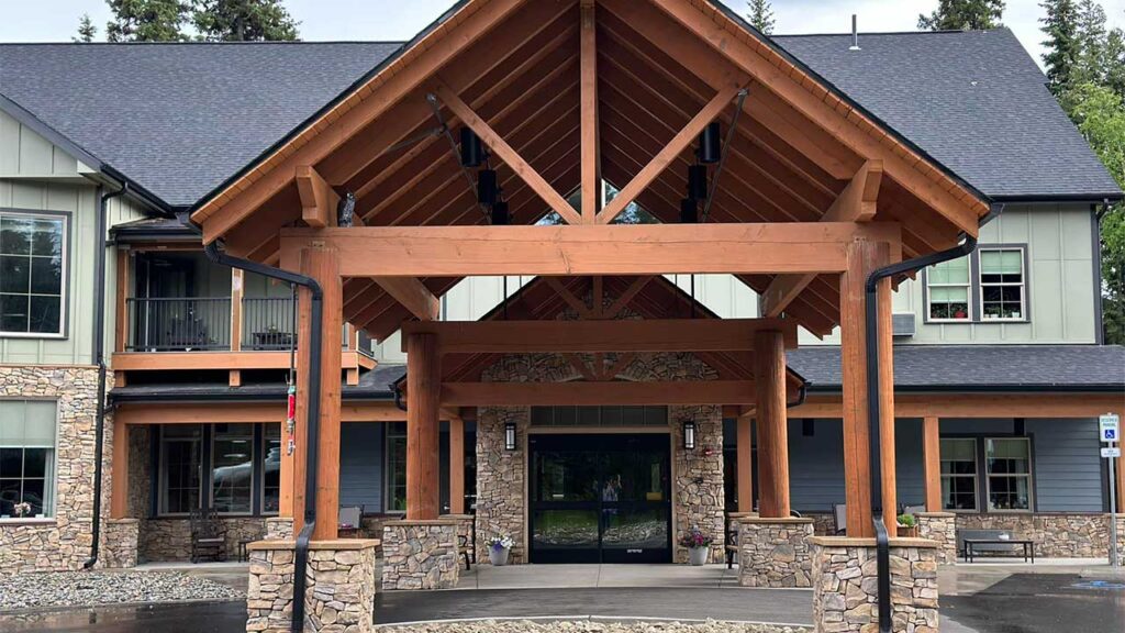 A large, rustic building with a stone base and wooden beams. It features a prominent covered entrance and large windows. The structure is surrounded by trees and has a paved driveway leading to the entrance.