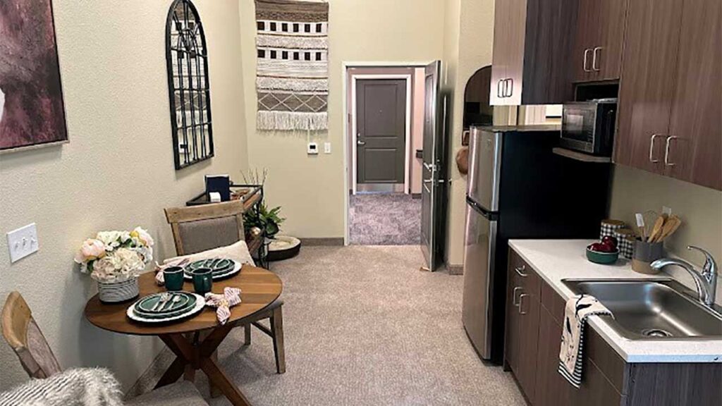 A compact kitchenette with dark wood cabinets, a small round dining table set with green plates, and a stainless steel fridge. The room is decorated with a woven wall hanging and has a neutral color scheme. The entry door is visible in the background.