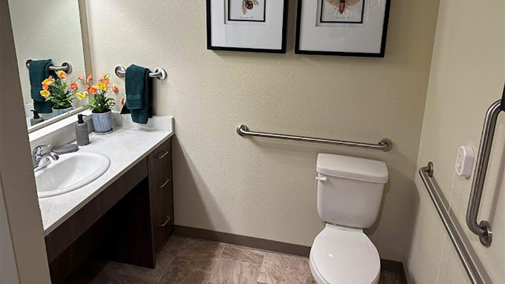 A bathroom with a white sink and toilet. The sink has a bouquet of flowers and a soap dispenser. Two framed pictures hang above the towel rack with green towels. The floor is tiled, and grab bars are installed near the toilet.