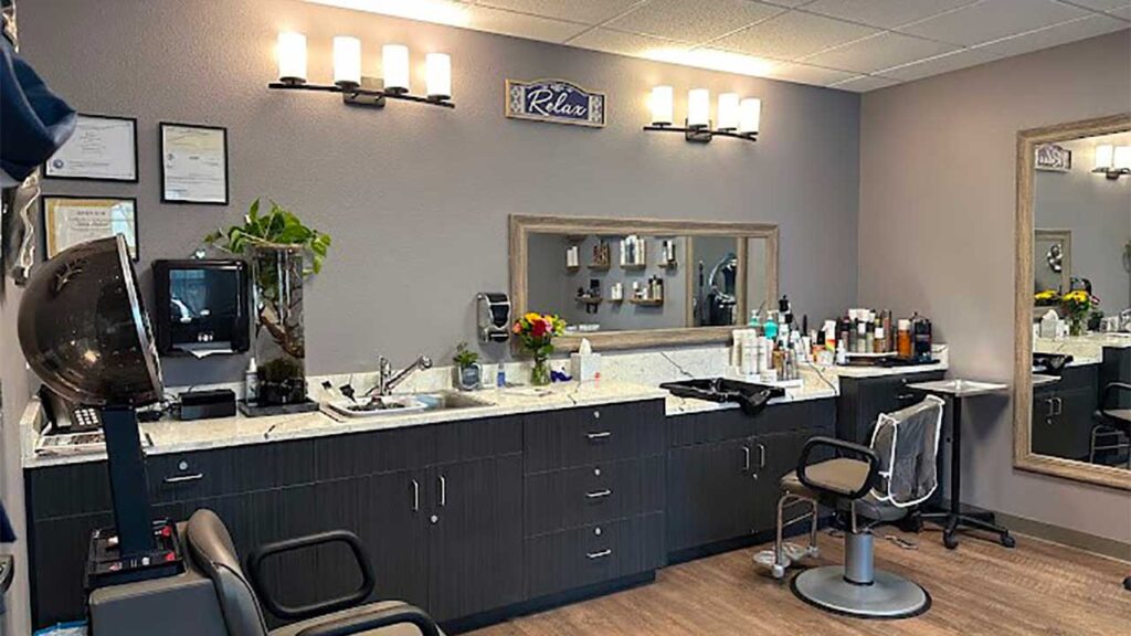 A cozy hair salon interior with a styling chair, large mirrors, and a counter filled with hair products. Warm lighting, plants, and a sign saying "Relax" create a welcoming atmosphere.