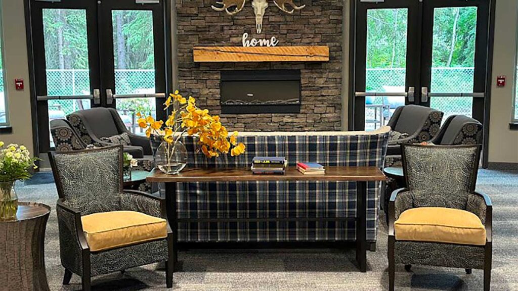 A cozy living room features a plaid sofa and two patterned armchairs with yellow cushions around a wooden table. A stone fireplace is adorned with a "home" sign and antler decor, with large windows offering views of greenery outside.