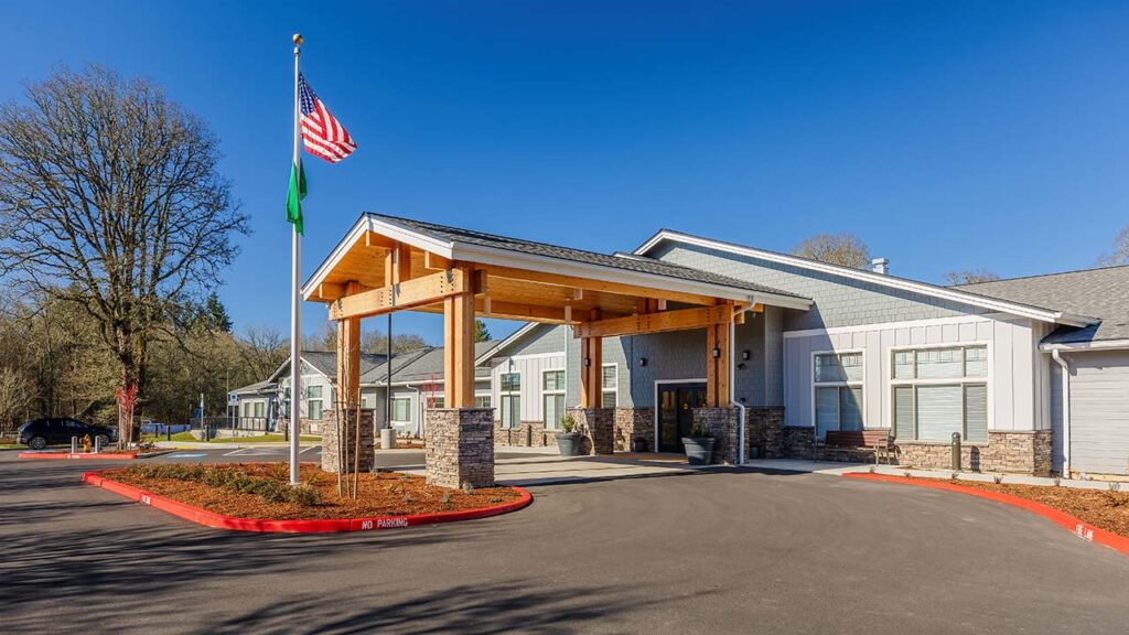 A modern building with a wooden entrance canopy and stone pillars. An American flag and another flag are flying on poles near the entrance. The building is surrounded by trees under a clear blue sky.