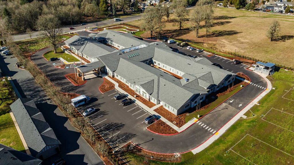 Aerial view of a large, single-story building with a gray roof, surrounded by a parking lot and landscaped areas. The structure, shaped like an H, is set in a suburban environment with trees and an open field nearby. Roads encircle the complex.