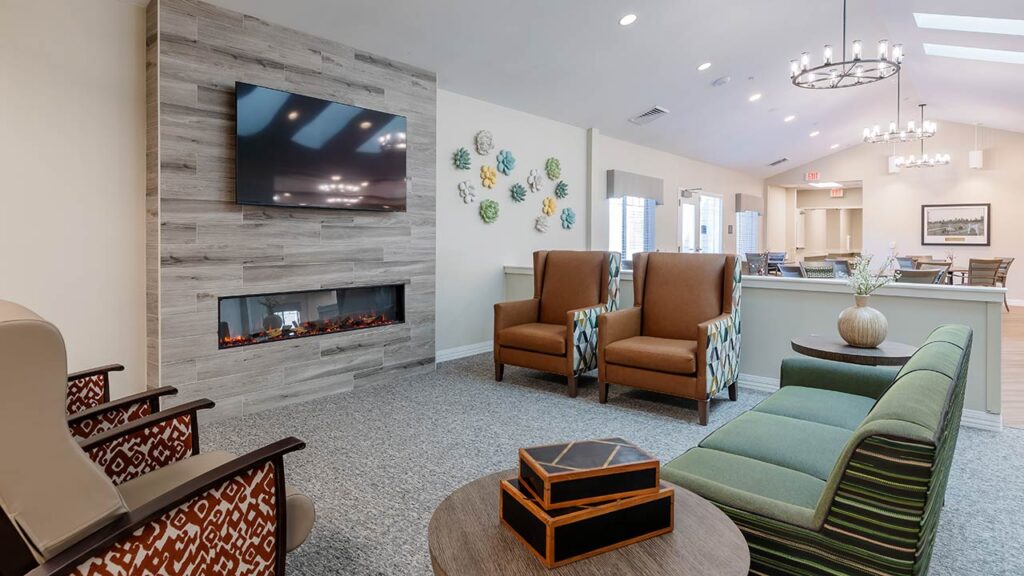 A modern communal living space with a wall-mounted TV above an electric fireplace, surrounded by wooden paneling. The room features two patterned chairs, a green-striped sofa, a round coffee table, and decorative wall art, with dining area visible in the background.
