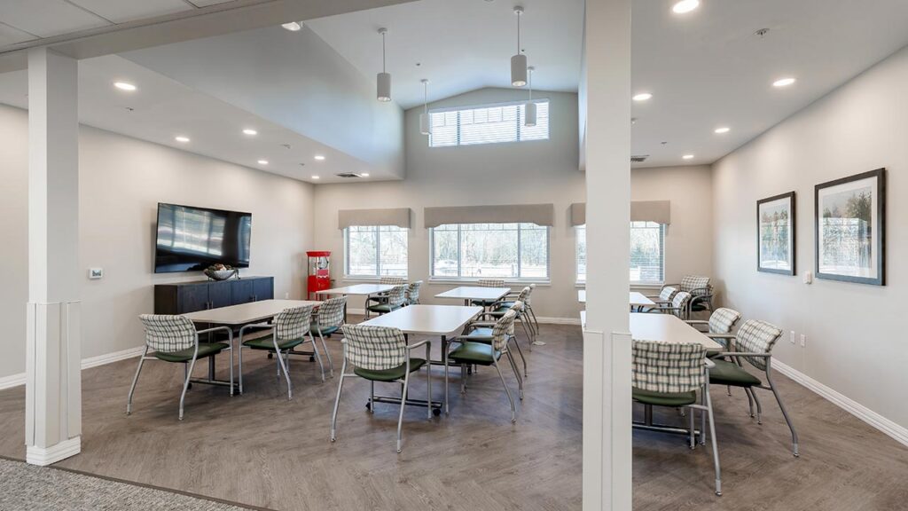 A spacious meeting room with several rectangular tables arranged in a T-shape. The room has cushioned chairs, a wall-mounted TV, large windows, and framed artwork. The flooring is wood, and the walls are light-colored. Ceiling lights illuminate the space.