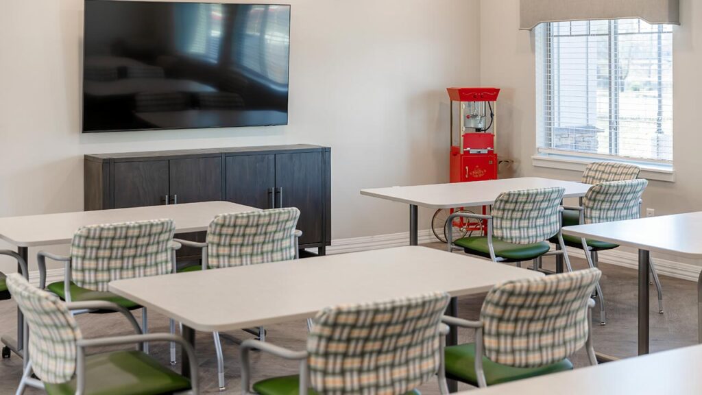 Classroom with white tables and padded chairs arranged in rows. A large flat-screen TV is mounted on a wall above a dark cabinet. There is a popcorn machine beside a window with blinds, letting in natural light.