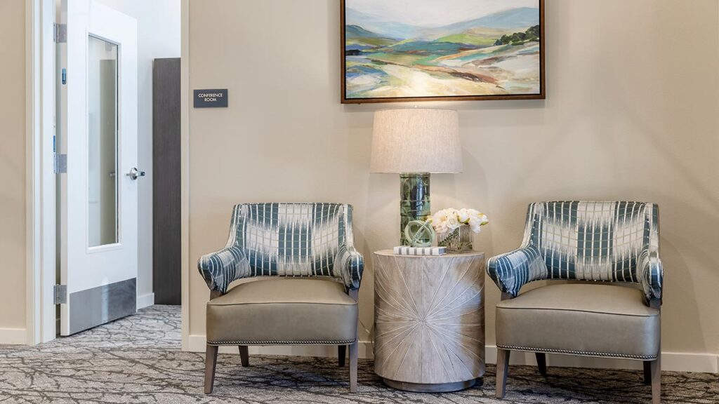 Two patterned armchairs flanking a round wooden table with a lamp and flowers, set beneath a landscape painting. An open door labeled "Conference Room" is partially visible to the left. Carpeted floor with a geometric pattern.