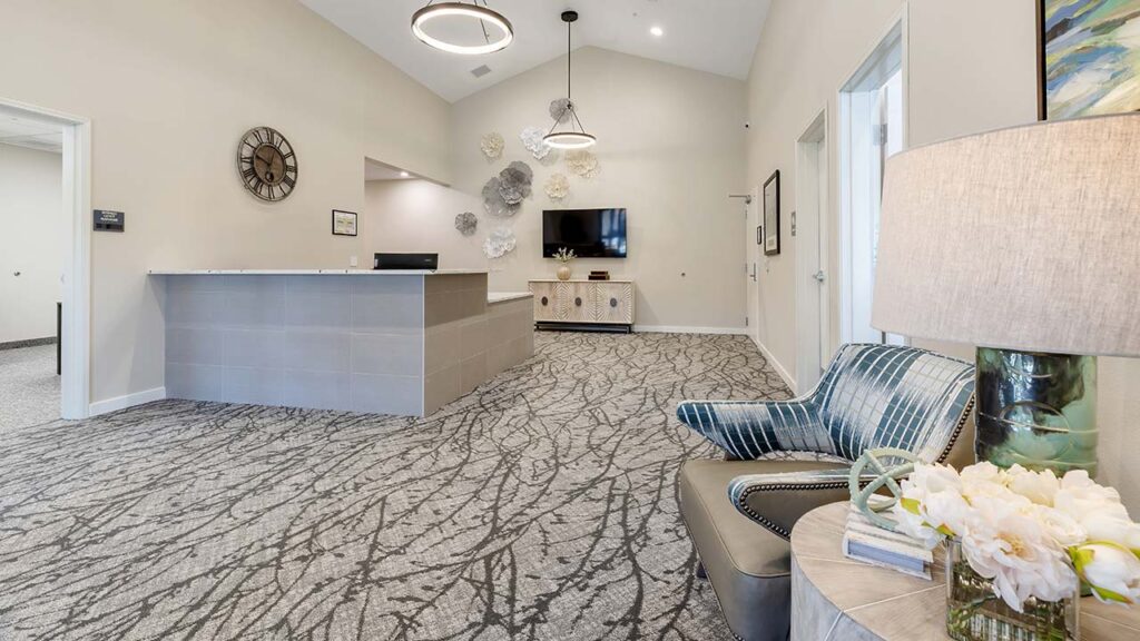 A modern waiting room with patterned carpet, a reception desk, decorative wall art, a large wall clock, and a flat-screen TV. There's a chair with a pillow and a side table with flowers next to a lamp. Neutral colors create a cozy atmosphere.
