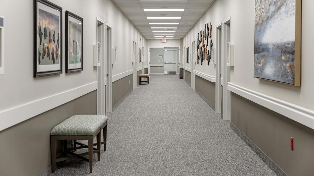 A long, empty hallway with gray carpet and beige walls, adorned with framed artwork. A patterned bench sits on the left side. Fluorescent lights line the ceiling, and a closed door is visible at the end of the corridor.