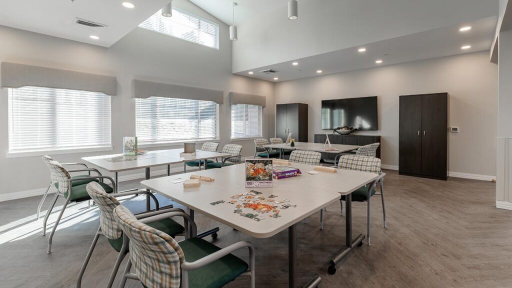 A well-lit room with large windows, several tables, and cushioned chairs. A partially completed puzzle and board games are on the tables. There's a TV and cabinets against a wall, with a light wood floor and recessed ceiling lights.