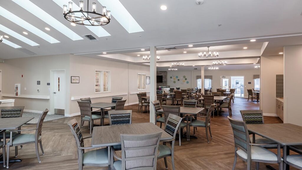 Spacious dining area with multiple wooden tables and chairs. The room features modern chandeliers, large windows, and white walls. The flooring is a mix of patterned and plain wood. There's a bright and inviting atmosphere.
