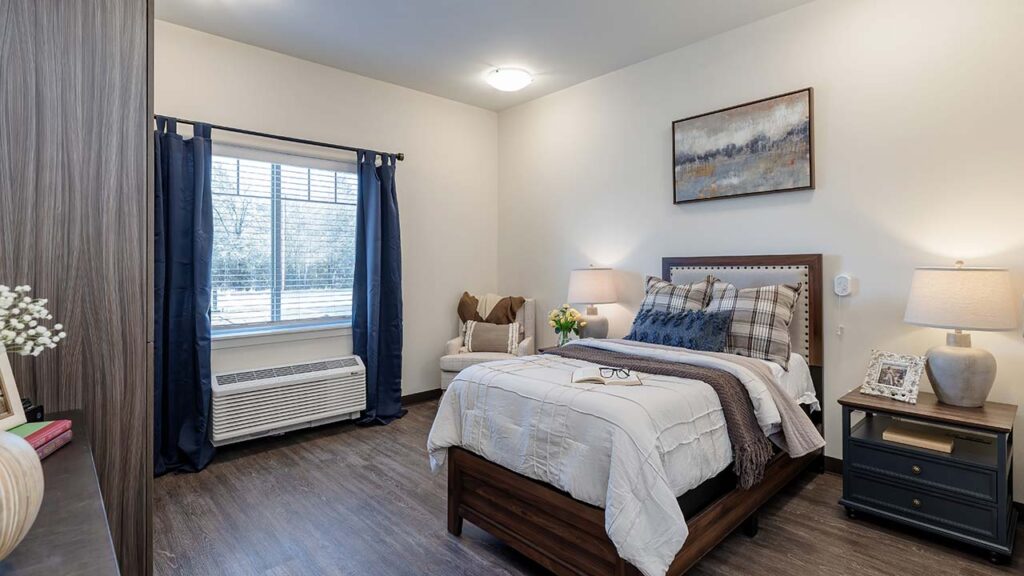 A cozy bedroom with a wooden bed, neatly made with a white comforter and plaid pillows. A bedside table with a lamp and picture frame is next to it. A chair and small table with flowers sit by the window with blue curtains. A landscape painting adorns the wall.