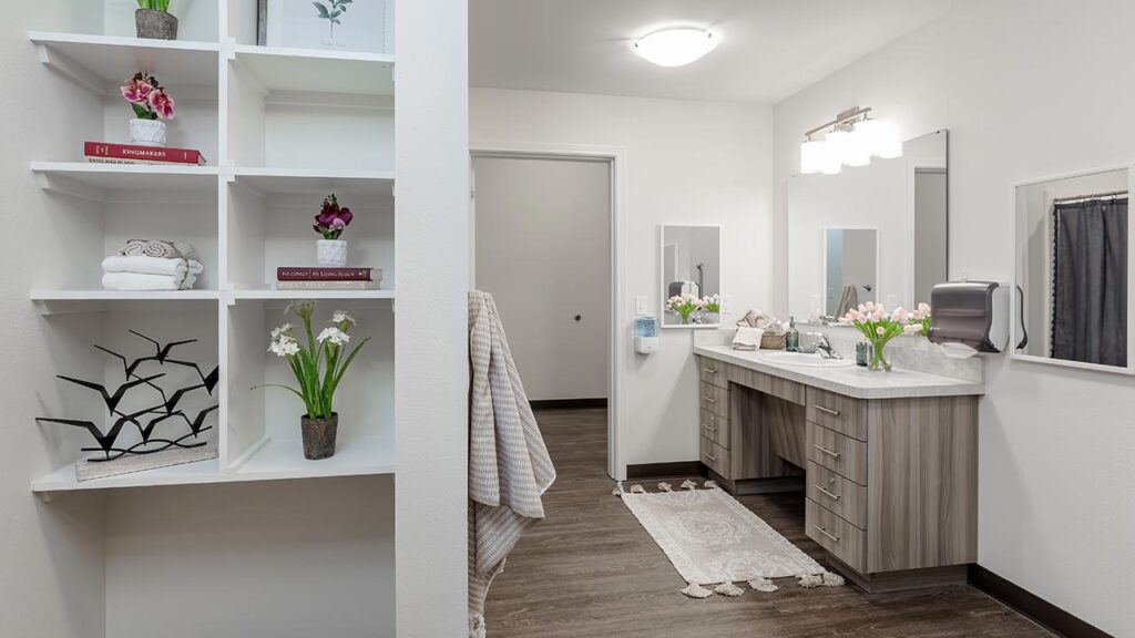 Modern bathroom with light wood cabinets, large mirror, and a decorative rug. Shelves display towels and small potted plants. The room features white walls, a wood floor, and soft overhead lighting.