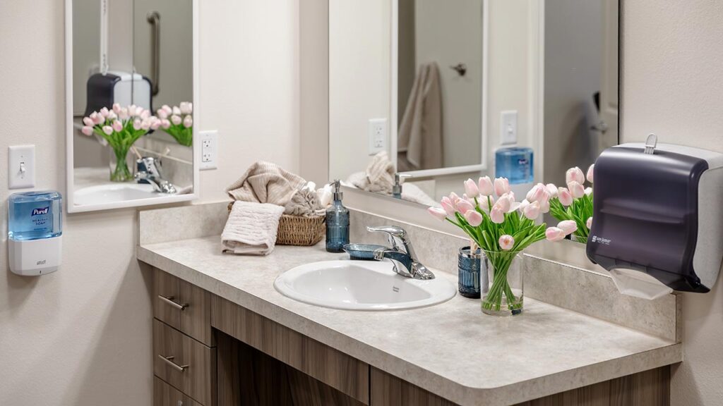 A bathroom counter with a sink, a faucet, and a vase of pink tulips. There are folded towels, a basket, a soap dispenser, and a paper towel holder. A large mirror reflects the flowers. The countertop has a light stone finish.