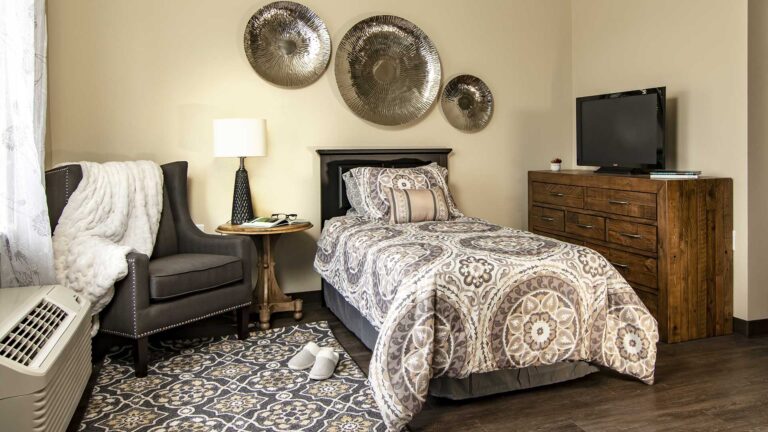 A cozy bedroom with a single bed featuring patterned bedding, a dark wooden dresser with a TV, a gray armchair with a white blanket, a small round table with a lamp, and metal wall art. A patterned rug and slippers are on the floor.