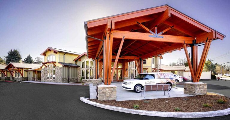 A modern building with a large wooden canopy entrance supported by beams. A white vehicle is parked under the canopy. The exterior features yellow walls, stone accents, and multiple windows. The sky is clear.