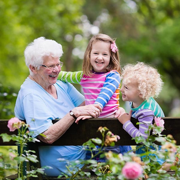 An elderly woman with white hair and a blue shirt is sitting on a bench surrounded by pink flowers, laughing with two young children in colorful striped shirts. They are outdoors in a green, leafy setting.