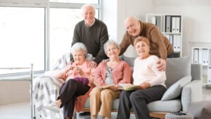 A group of five smiling elderly people, three women and two men, sitting and standing around a couch in a well-lit room. Two women are holding books, and a walker is visible beside the couch. Shelves with binders are in the background.