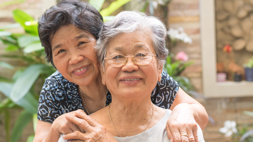 Two elderly women smile warmly at the camera in an outdoor setting with green plants in the background. One woman with short hair and glasses stands in front, while the other embraces her from behind, both wearing casual clothing.