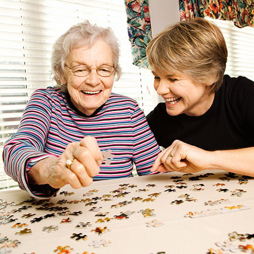 Two women sitting at a table, smiling and working on a jigsaw puzzle together. The older woman, wearing glasses and a striped shirt, holds a puzzle piece. The younger woman, in a black shirt, leans in, smiling. Bright light comes from nearby windows.