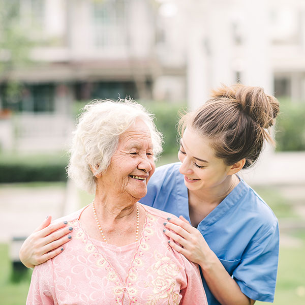 A smiling elderly woman in a pink sweater is outdoors, being gently embraced by a young woman in blue scrubs. They are in a garden setting with blurred trees and buildings in the background, sharing a joyful moment.