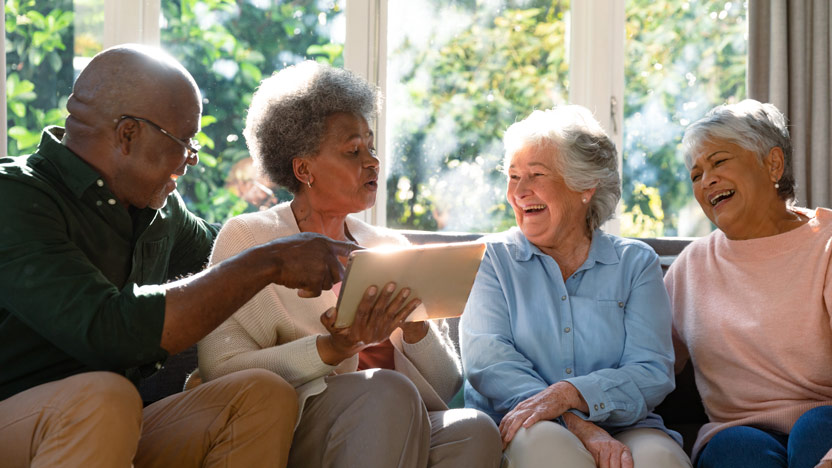 A group of four older adults sitting on a couch, smiling and laughing. One man is pointing at a tablet held by one of the women. Sunlight streams through a window behind them, creating a warm and cheerful atmosphere.