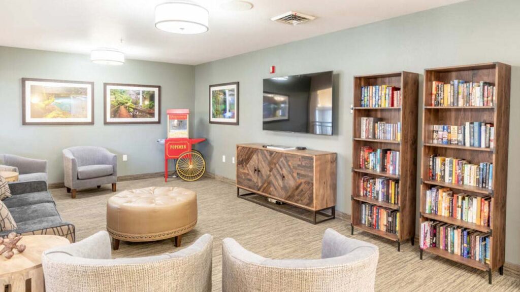 A cozy room with gray walls features a TV mounted on the wall, flanked by bookshelves filled with books. A wooden cabinet sits below the TV. There are two armchairs, a circular ottoman, a popcorn machine, and framed pictures adorn the walls.