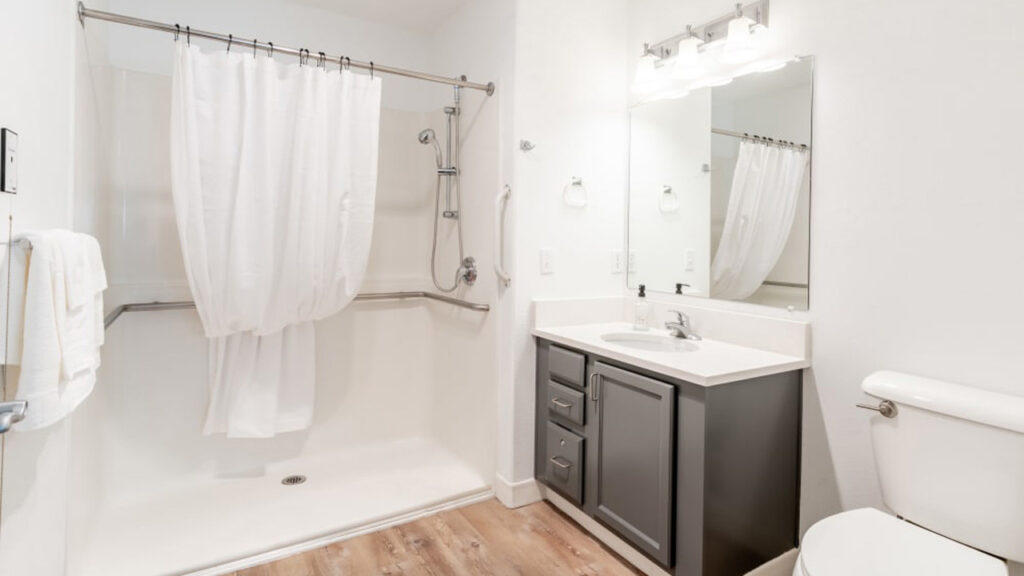 A modern bathroom featuring a walk-in shower with a white curtain, a dark vanity with a white countertop, a large mirror with lights above, a towel on a rack, and a white toilet. The floor is wood-textured.