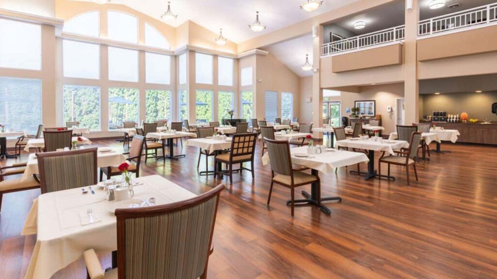 A spacious and elegant dining area with wooden floors and large windows. The room features multiple tables with white tablecloths and chairs. Natural light floods the space, highlighting a high ceiling and soft, neutral-toned walls.