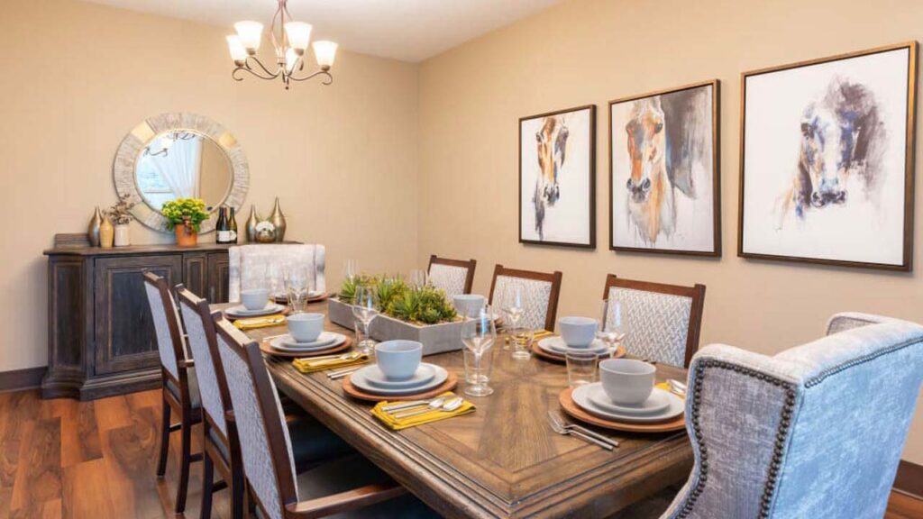 A dining room with a wooden table set for eight, featuring gray chairs, plates, and glasses. A centerpiece with greenery is on the table. The room has beige walls, three abstract horse paintings, a circular mirror, and a chandelier.