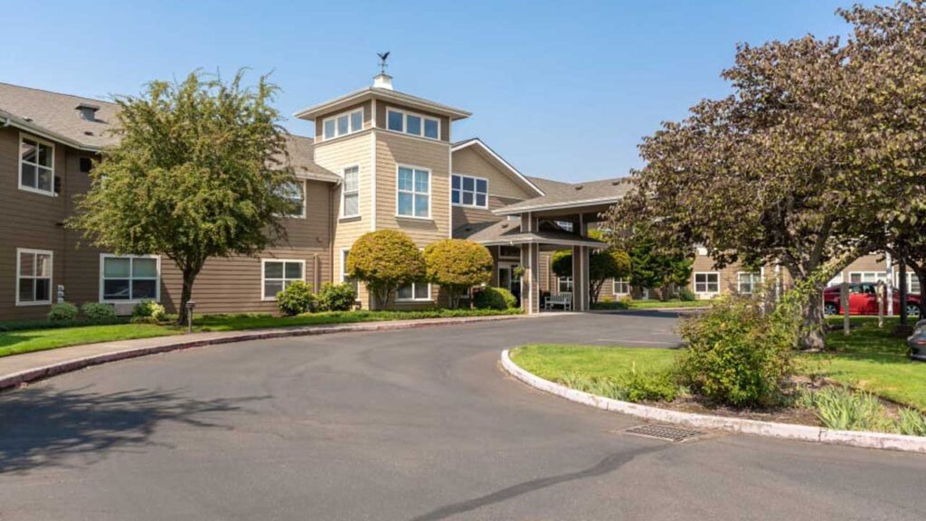 A two-story building with a light brown exterior, surrounded by trees and shrubs. There's a driveway leading to a covered entrance, and the area is sunny with a clear blue sky.