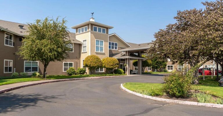 A two-story beige building with large windows, surrounded by trees and a paved driveway. The entrance features a covered walkway. The setting appears peaceful with neatly trimmed grass and a clear blue sky.