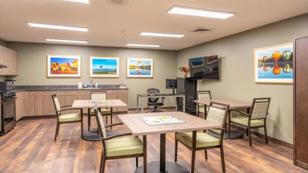 A communal room with wooden floors and beige walls features several tables and chairs. Colorful hot air balloon artwork adorns the walls. A black TV is mounted nearby, and a kitchen area with cabinets is in the background.