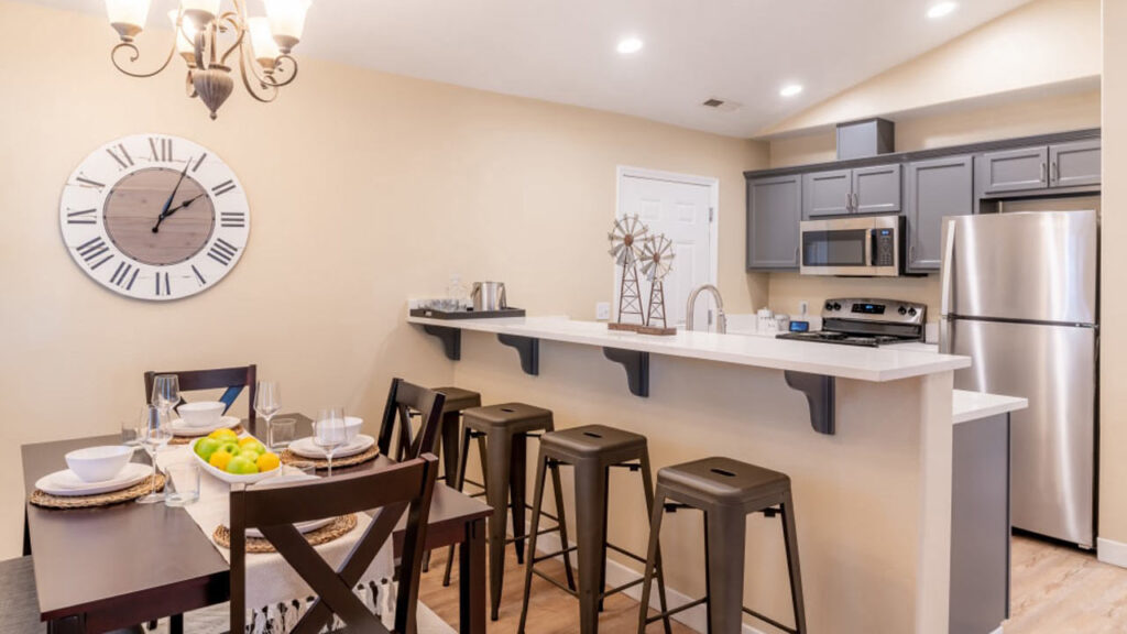 A modern kitchen and dining area featuring a wooden table set for four, metal bar stools at a white counter, gray cabinets, stainless steel appliances, and a large clock on a beige wall. A decorative windmill piece sits on the counter.