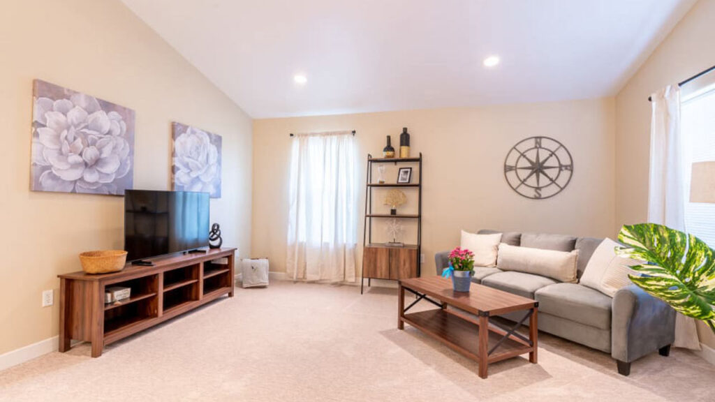 A cozy living room with beige walls features a gray sofa, a wooden coffee table, and a large TV on a stand. Two floral paintings hang on the wall. A window with sheer curtains allows natural light. A plant sits next to the sofa, adding a touch of greenery.