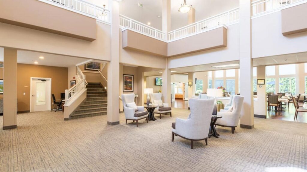 Spacious, well-lit lobby with high ceilings, a staircase, and seating area featuring cushioned chairs and small tables. Neutral colors dominate the decor. Large windows provide natural light, and the second floor is visible with white railings.