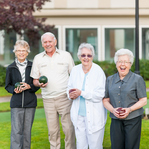 Four elderly people are smiling and holding bocce balls outdoors on a lawn. They appear to be enjoying a game together, standing in front of a building and trees in the background.