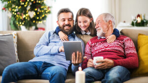 Three people sitting on a couch are smiling at a tablet. A young girl is hugging a bearded man, who is showing her the screen. An older man in a red sweater is holding a mug. A Christmas tree is in the background.