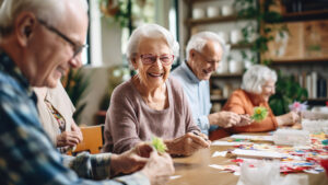 A group of elderly people smiling and crafting colorful paper flowers at a table in a bright room with plants. They appear engaged and happy, focusing on their creative activity together.