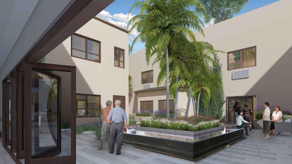 A group of people stand and sit in a modern courtyard surrounded by beige buildings. A tall palm tree and plants are in the center. Large windows and glass doors are visible, with natural light illuminating the space.