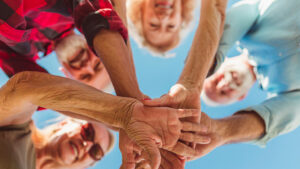 A group of four people forming a circle overhead, stacking their hands together in the center. They are smiling and looking down towards the camera, with a clear blue sky in the background.