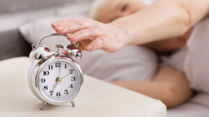 A person in bed reaches out to touch a silver analog alarm clock on a bedside table. The clock shows 7:00. The scene suggests waking up in the morning.