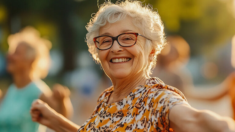 Smiling elderly woman with short gray hair and glasses, wearing a patterned shirt, enjoying herself outdoors in warm sunlight, with blurred people and greenery in the background.