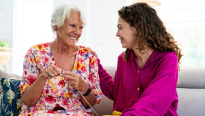 An older woman and a younger woman sit together on a couch, smiling warmly at each other. The older woman wears a floral dress and knits, while the younger woman, in a bright pink blouse, rests her hand on the other’s shoulder.