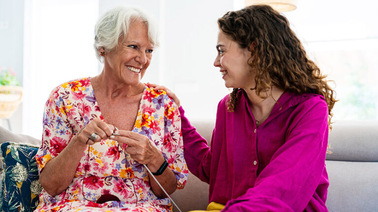 An older woman and a younger woman sit together on a couch, smiling warmly at each other. The older woman wears a floral dress and knits, while the younger woman, in a bright pink blouse, rests her hand on the other’s shoulder.